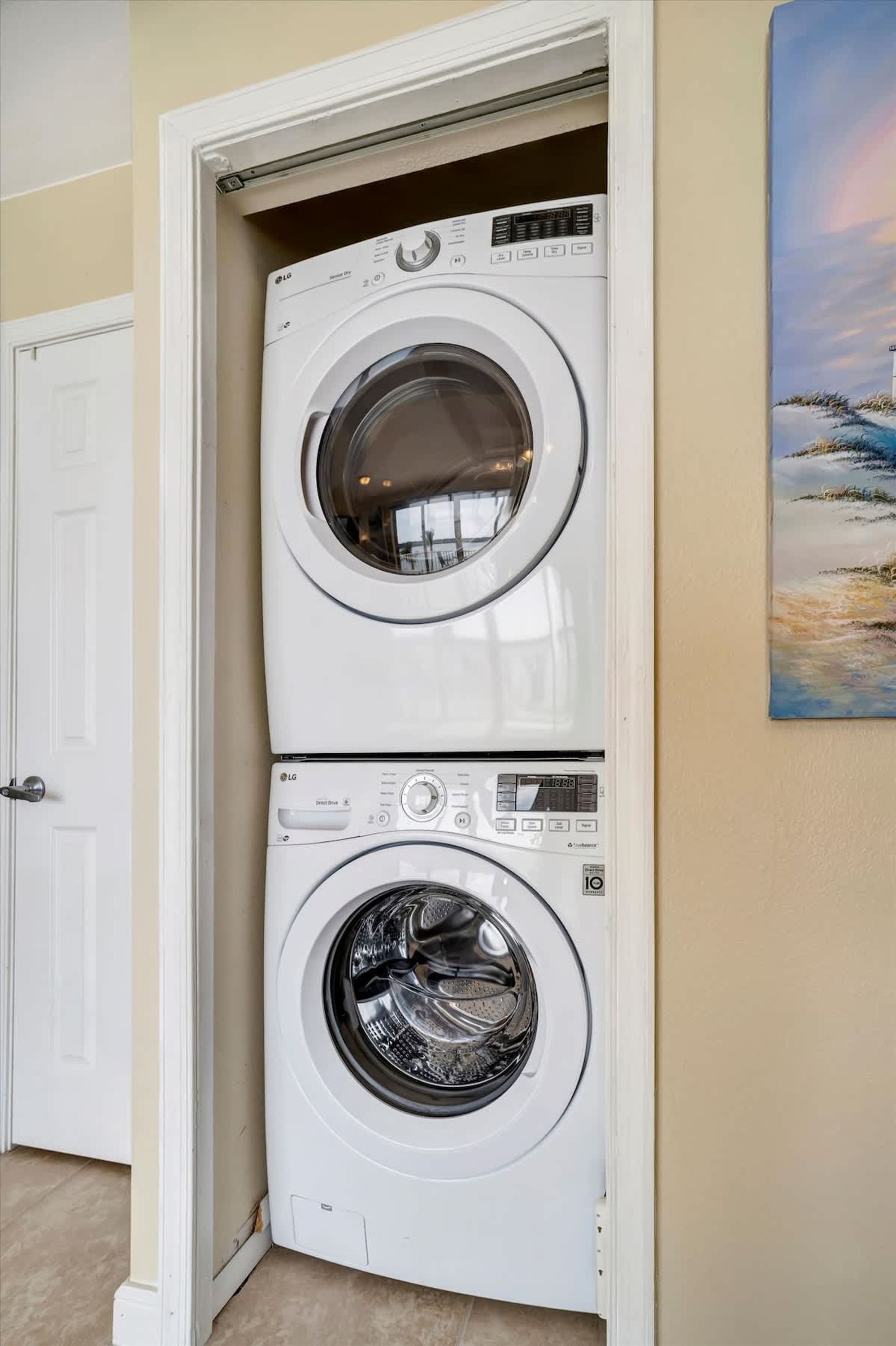 A stacked washer and dryer unit in a corner with a door and a painting on the wall.