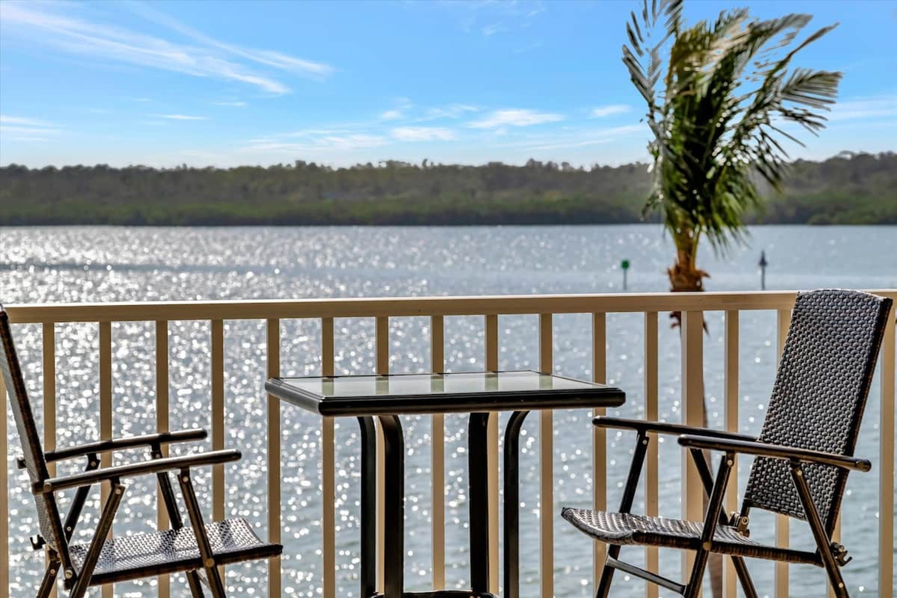 A balcony with a glass table and two chairs, overlooking a body of water with a palm tree and a forested shoreline.