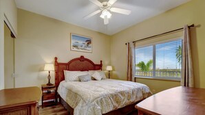 A bedroom with a bed, two bedside tables with lamps, a ceiling fan, a window with blinds, and a painting on the wall.