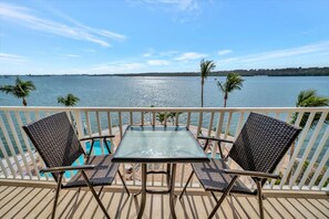 A balcony with a glass table and two wicker chairs, overlooking a body of water with palm trees and a distant shoreline. - Sunlit Bayview Suite Balcony Pool Hot Tub (St. Petersburg)