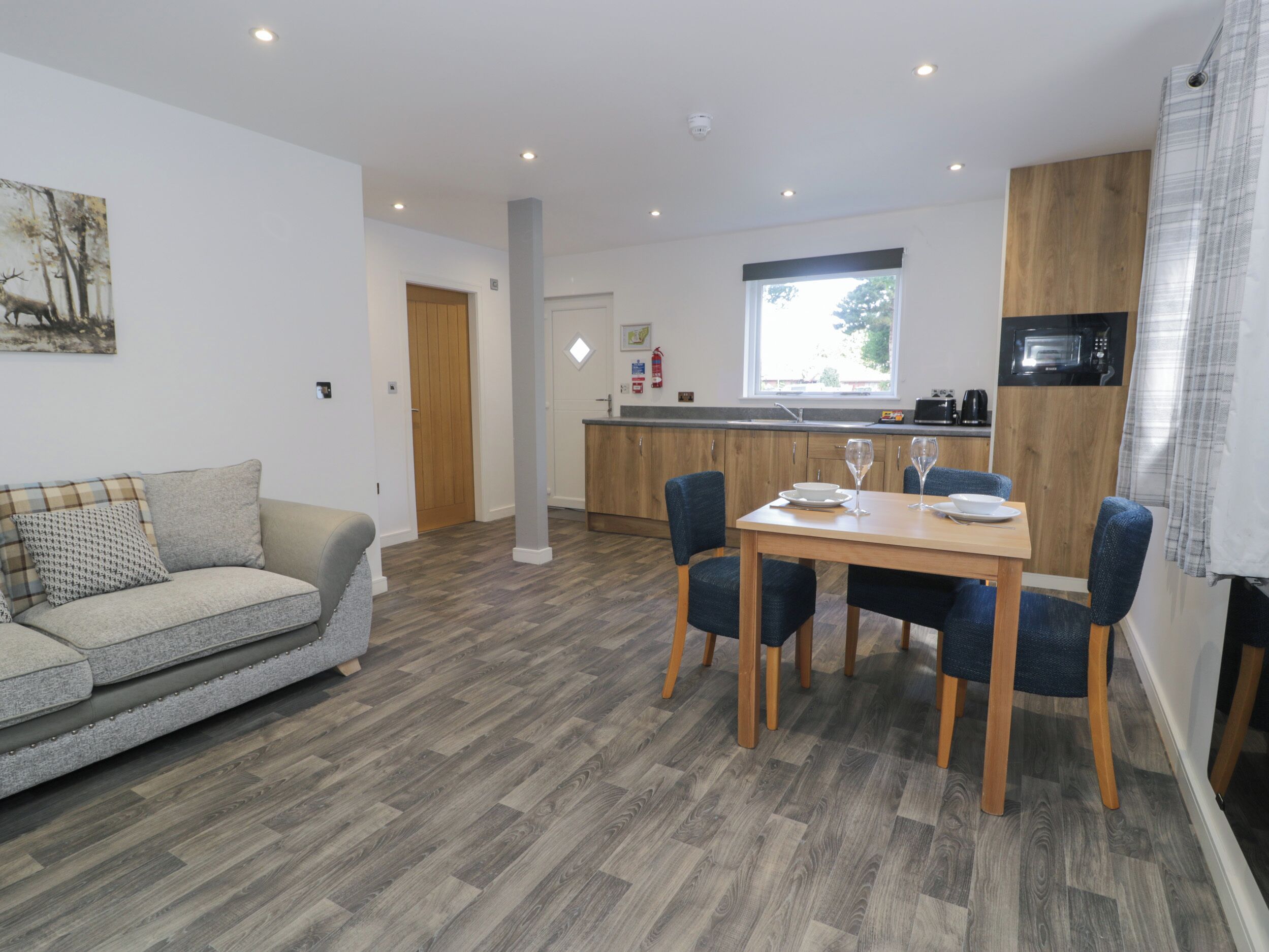 A modern living room with a sofa, a dining table with chairs, a kitchen area with a microwave, and a window with a view of trees.