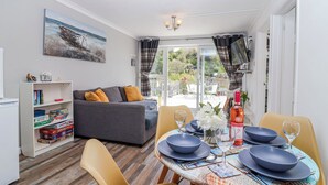 A cozy living area with a sofa, dining table set for four, and a bookshelf with children's books. - Shells Cottage (Looe)