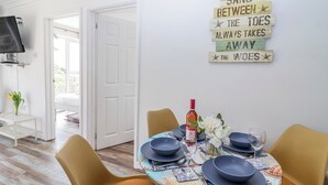 A dining area with a table set for four, a bottle of wine, and a decorative sign on the wall. - Shells Cottage (Looe)