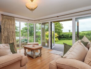 A room with a wooden table, two sofas, a large window with a view of a garden, and a ceiling light. - Hole Park (Holsworthy)