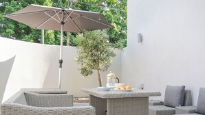 Un área de patio al aire libre con mobiliario de mimbre, una mesa con una cafetera y un paraguas grande.