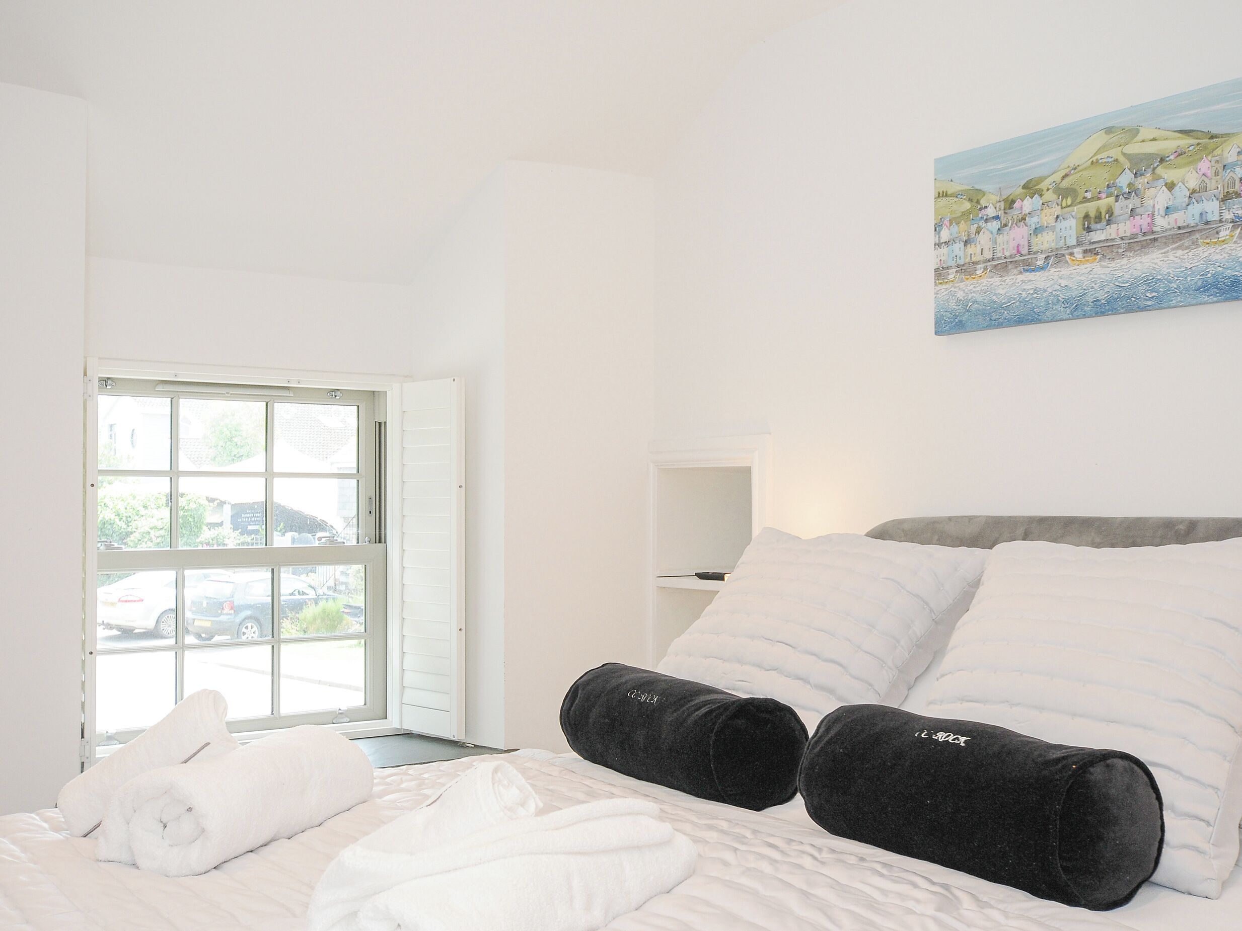 Ein Hotelzimmer mit Bett, weißer Bettwäsche, einem Bild an der Wand und einem Fenster mit Blick auf Autos und Grünflächen.