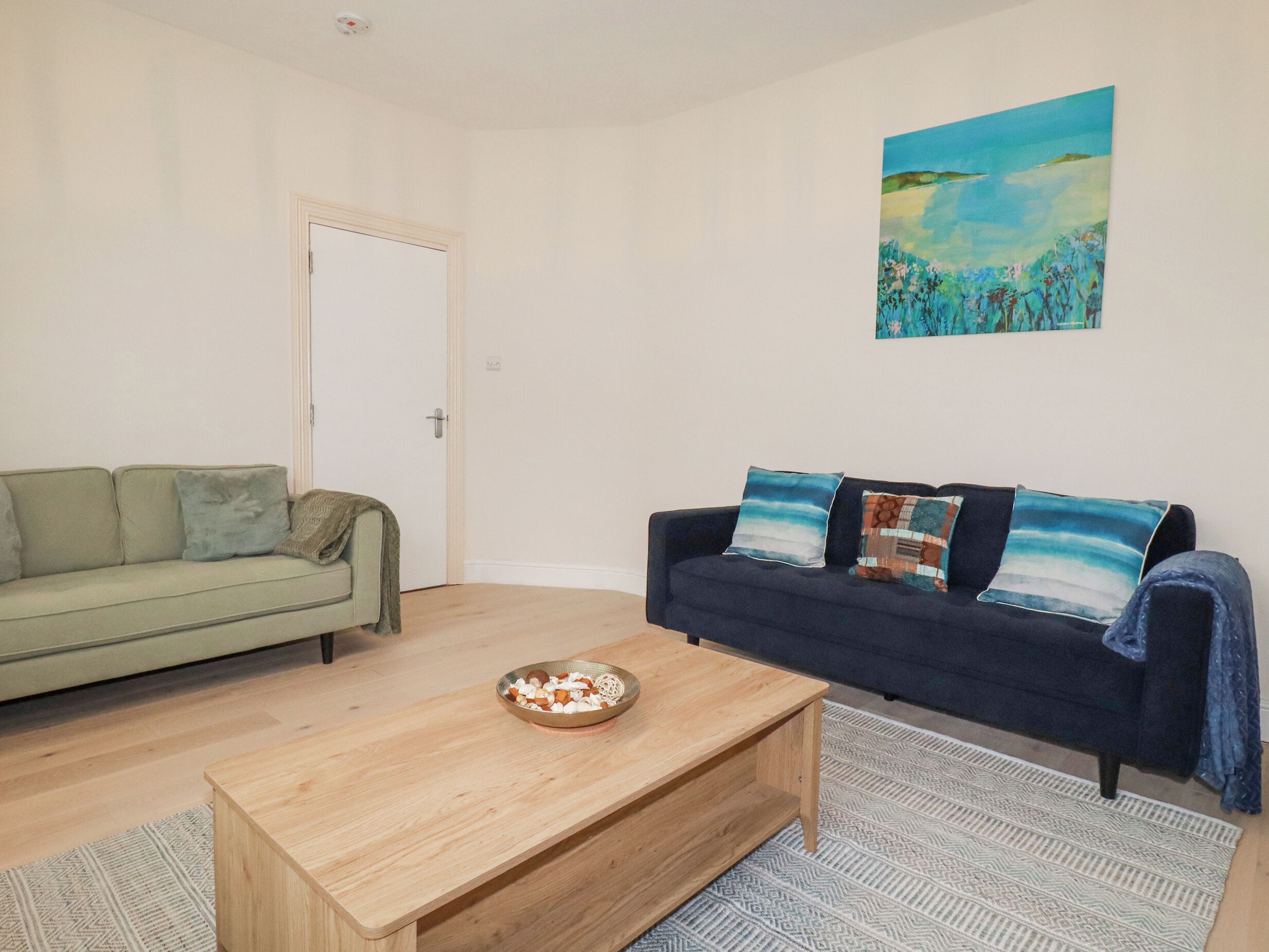 Een zithoek met een lichtgroene bank, een donkerblauwe bank met gekleurde kussens, een houten salontafel en een schilderij aan de muur.