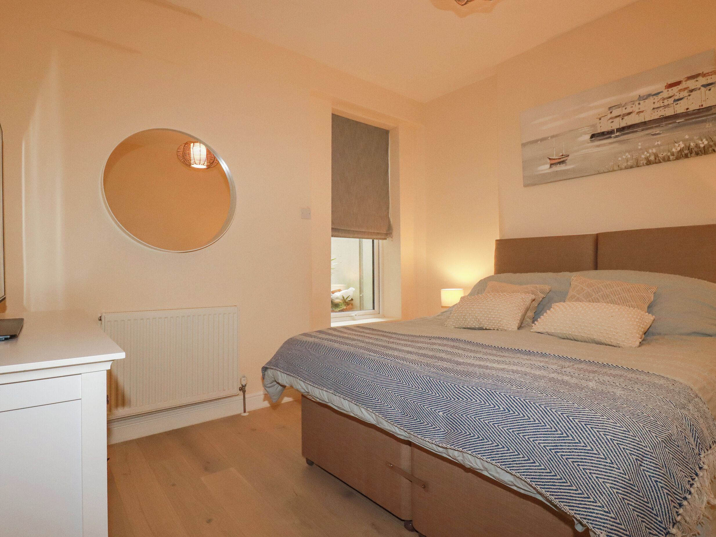 A bedroom with a bed, a round mirror, a radiator, a window with a view, and a painting on the wall.