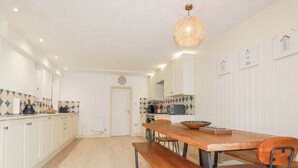A modern kitchen with white cabinets, a wooden dining table with benches, and a hanging light fixture. - Serendib Cottage (Newquay)