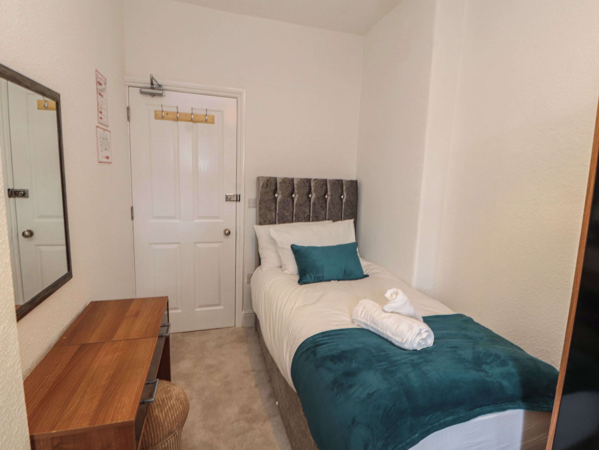 A small, neatly arranged bedroom with a single bed, a wooden desk, and a door with a coat rack.