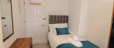 A small, neatly arranged bedroom with a single bed, a wooden desk, and a door with a coat rack.