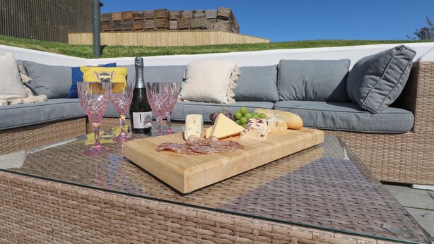 An outdoor seating area with a wicker table, a bottle of wine, glasses, cheese, and grapes. - Piglet Cottage (Haverfordwest)