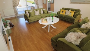 A spacious living room with two green sofas, a white coffee table, and a large window with curtains. - Piglet Cottage (Haverfordwest)