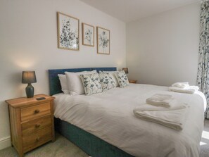A neatly made bed with white linens and floral-patterned pillows, a wooden nightstand with a lamp, and two framed floral artworks on the wall. - Dalegarth Hall Farm Cottage 2 (Holmrook)