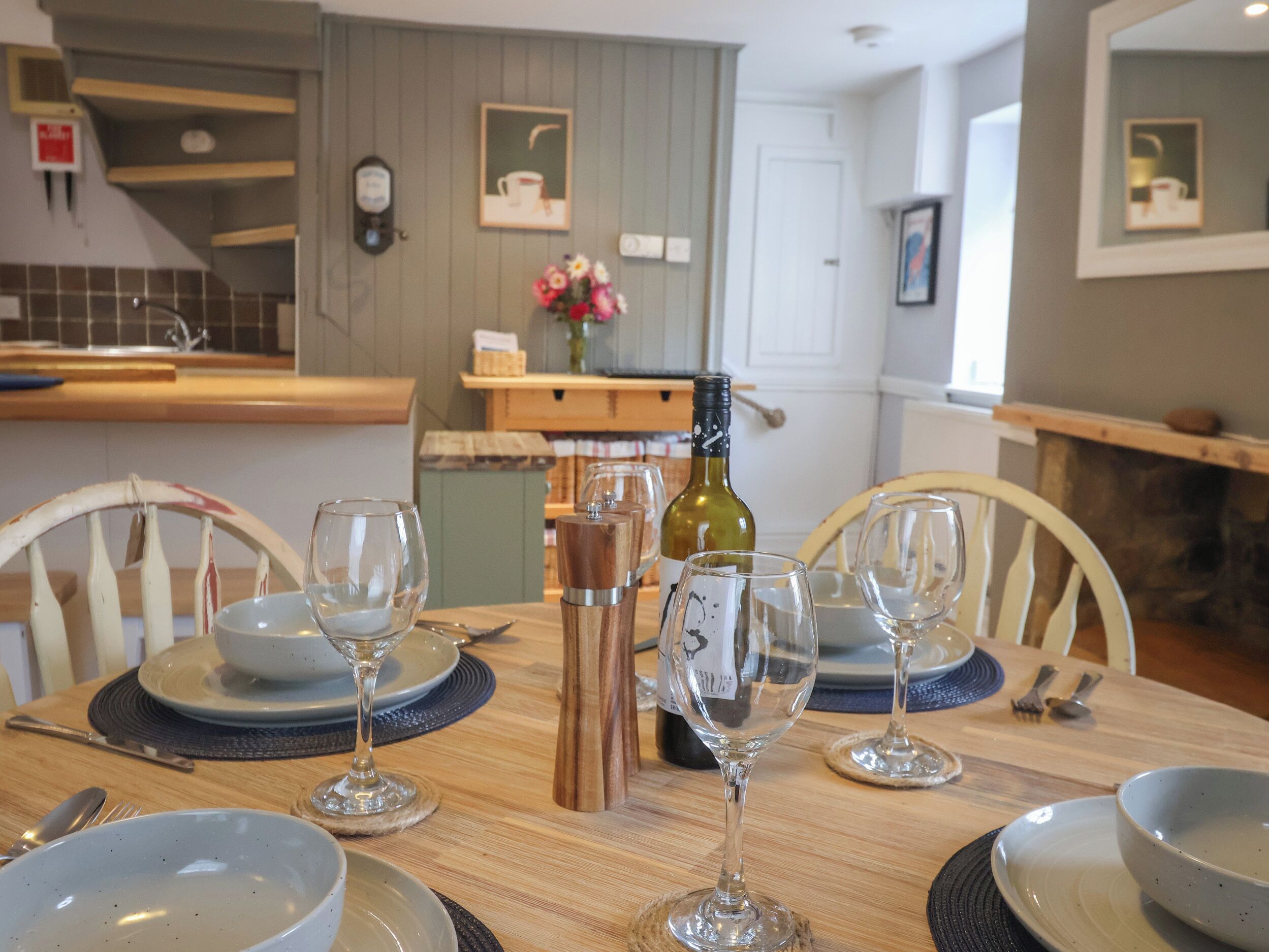 A dining area with a wooden table set for four, surrounded by white chairs. A bottle of wine and glasses are on the table.
