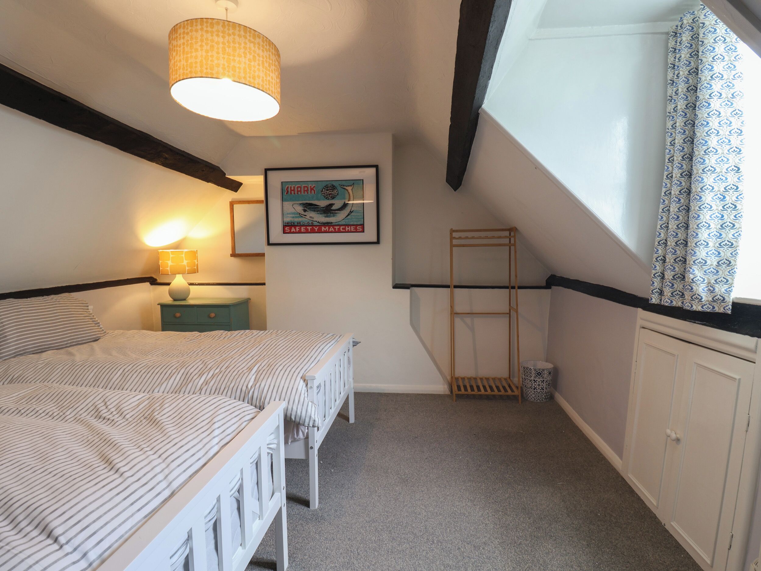 A bedroom with two beds, a hanging light fixture, a framed poster, a window with patterned curtains, and a small wooden shelf.