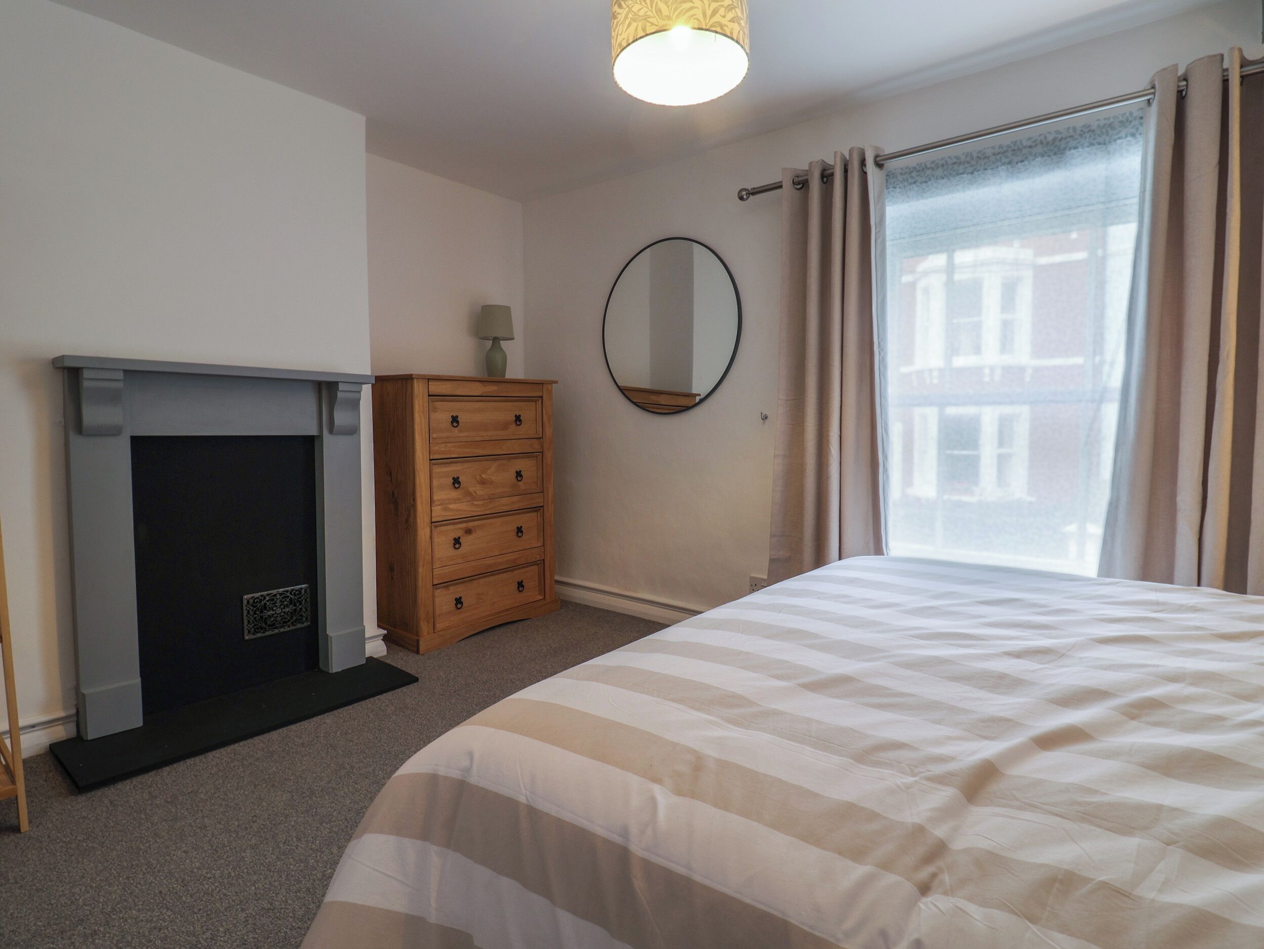 A bedroom with a bed, a dresser, a mirror, a fireplace, and a window with curtains.