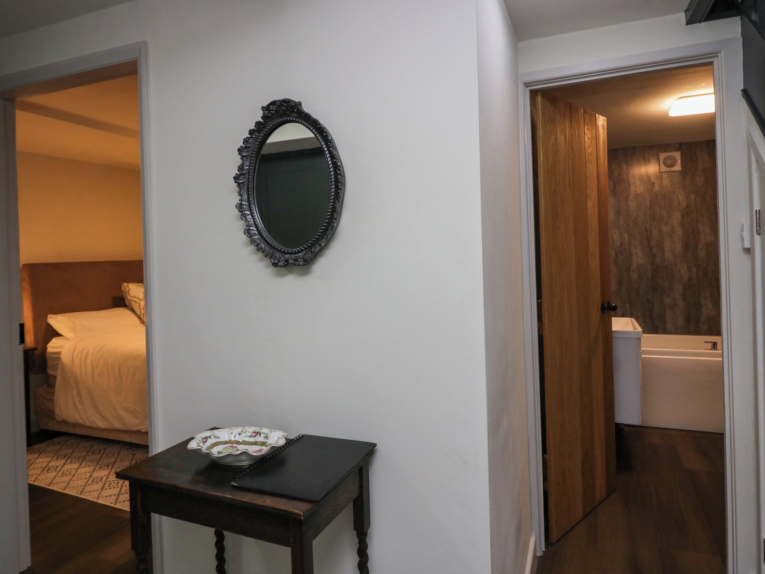 Una habitació d'hotel amb un llit, una taula de nit amb un bol decoratiu, un mirall i un bany visible a través d'una porta oberta.