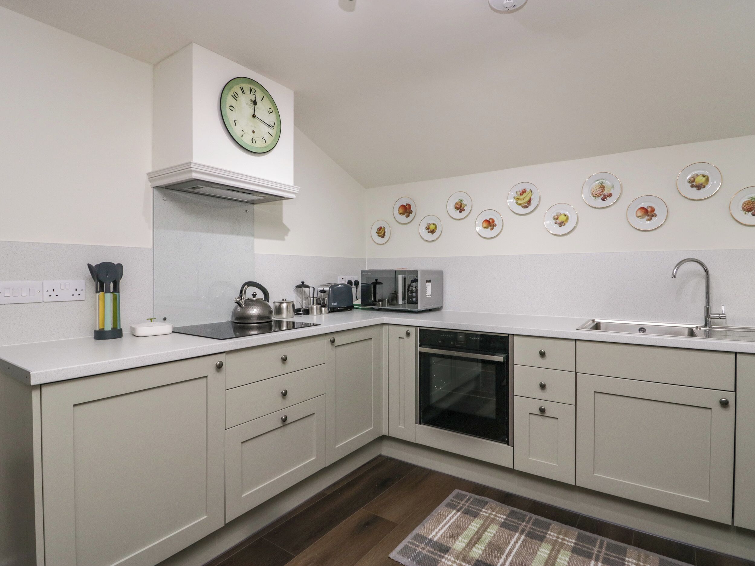 A modern kitchen with white cabinets, a built-in oven, a microwave, and a sink. There are wall-mounted plates with fruit designs and a clock above the stove.