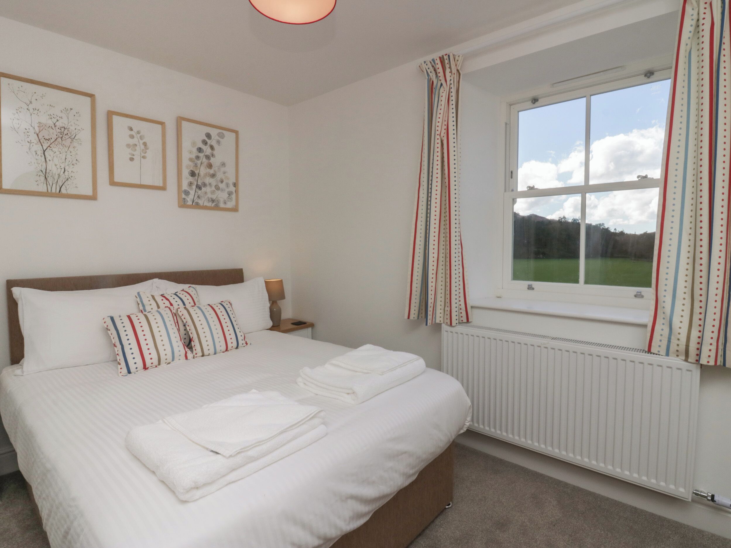 A neatly made bed with white linens and striped pillows, a bedside table with a lamp, and a window with curtains.