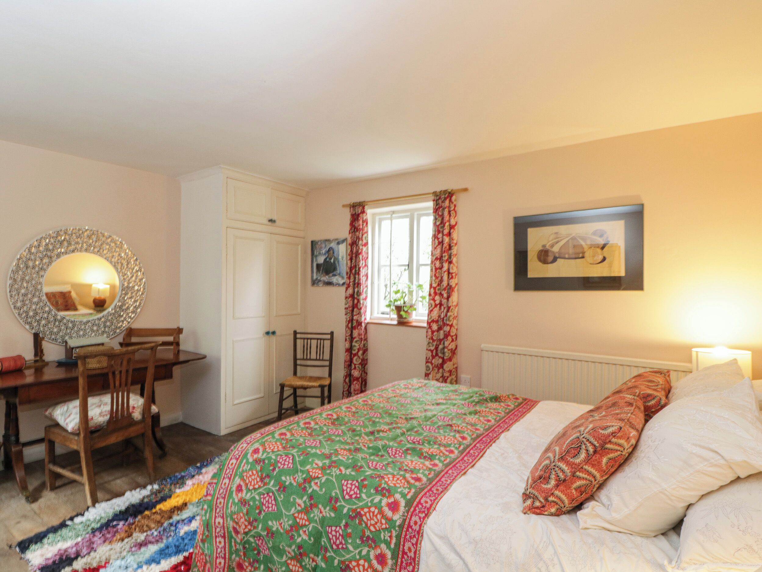 A bedroom with a bed, a chair, a desk, a mirror, a window with curtains, and a radiator.