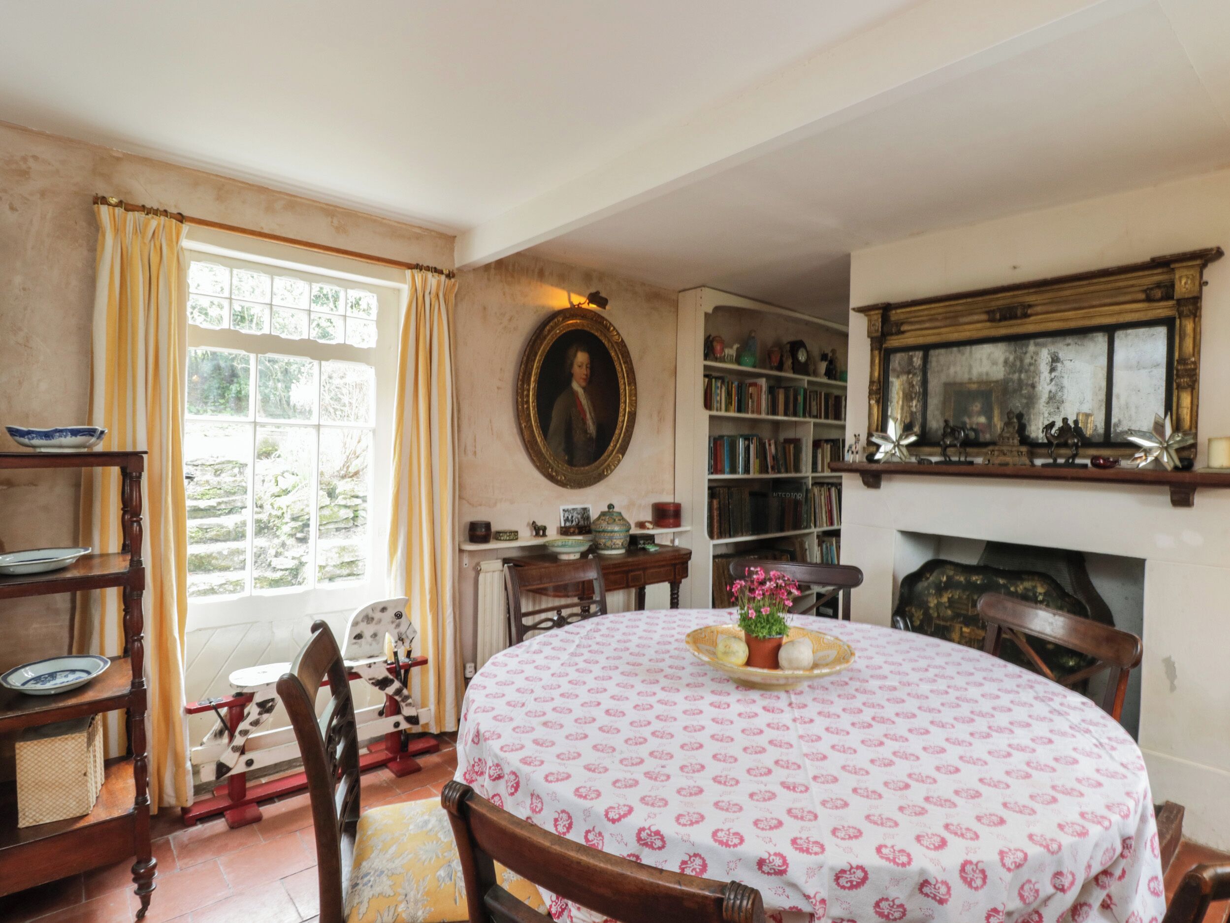 A traditional dining room with a round table, chairs, a fireplace, bookshelves, and a window with curtains.