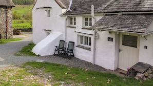 A white cottage with a stone chimney, a small porch with two chairs, and a gravel driveway.