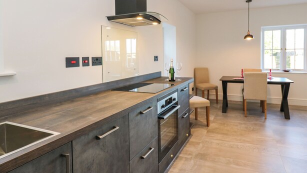 A modern kitchen with a large island, built-in oven, and a dining area with a table and chairs. - Luham Cottage (Penrith)