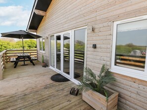 Una terraza de madera con mesa de picnic y sombrilla, una puerta corrediza de vidrio y vistas al campo.