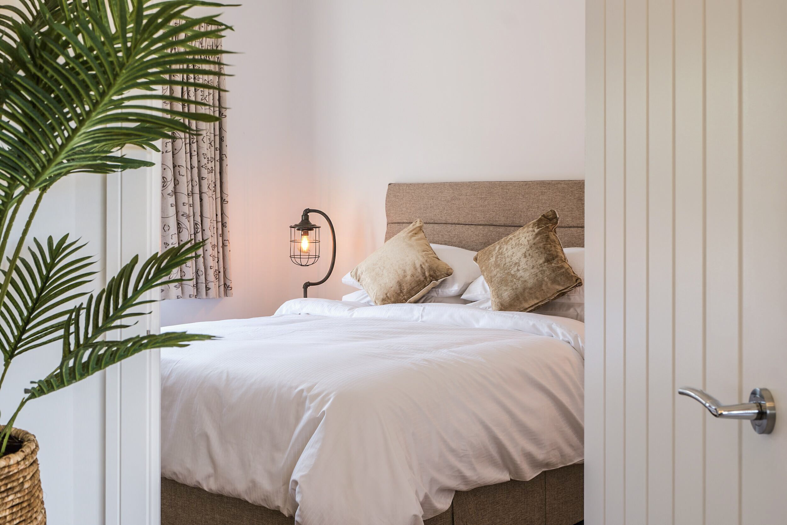 Een net opgemaakt bed met witte lakens en twee kussens, een bedlampje en een plant in pot.