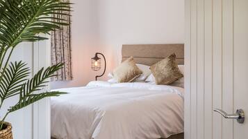 Een net opgemaakt bed met witte lakens en twee kussens, een bedlampje en een plant in pot.