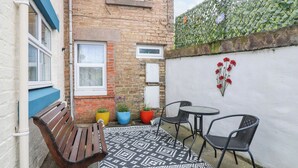 Un petit patio extérieur avec un banc en bois, une table ronde et quatre chaises. On y trouve des plantes en pot et un tapis à motifs sur le sol.