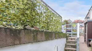 Un mur blanc surmonté d’une clôture métallique recouverte de feuilles vertes, un escalier en pierre et un petit espace aménagé en jardin.
