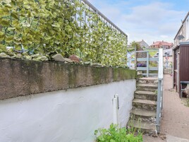Una pared blanca con una cerca de metal cubierta de hojas verdes, una escalera de piedra y un pequeño jardín.