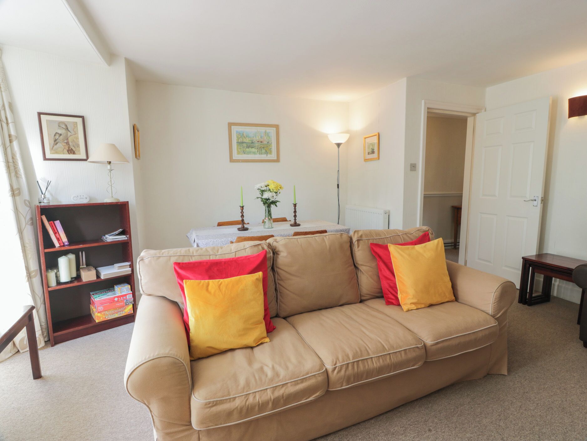 A living room with a beige sofa, two red and yellow cushions, a bookshelf, a table with a vase of flowers, and framed pictures on the wall.