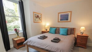 Une chambre à coucher bien rangée, avec un lit, deux tables de chevet, une fenêtre avec des rideaux et deux tableaux encadrés accrochés au mur.