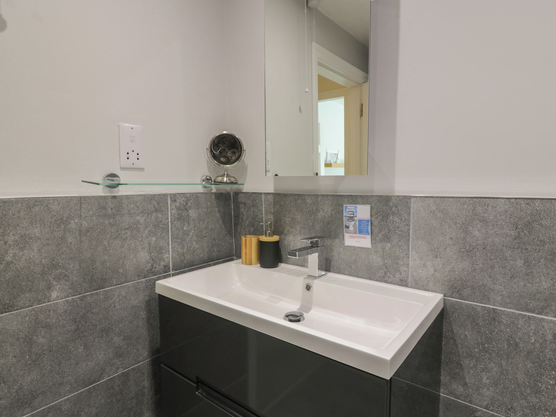 A modern bathroom with a white sink, dark tiles, a glass shelf with toiletries, and a mirror.