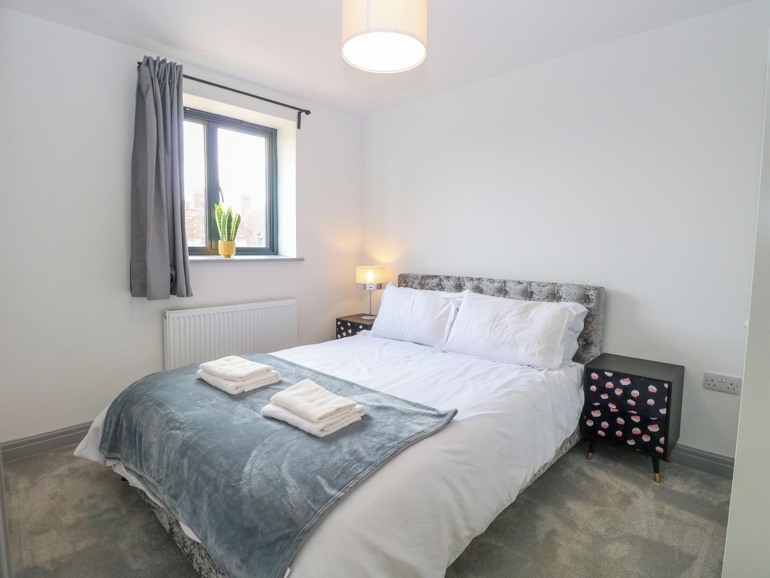 Cama bem arrumada com lençóis brancos e um cobertor azul, mesa de cabeceira com abajur, janela com cortinas e um radiador.