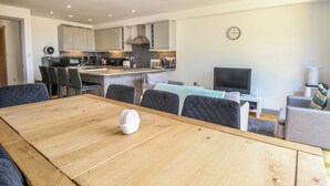 A modern kitchen with a central island, dining area with a wooden table and chairs, a television, and a clock on the wall. - Henham 3 Bernaleen Cottages (King's Lynn)