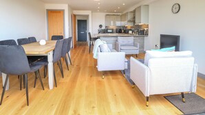 A modern kitchen with a central island, a dining area with a wooden table and chairs, and a living area with a sofa and armchairs. - Henham 3 Bernaleen Cottages (King's Lynn)