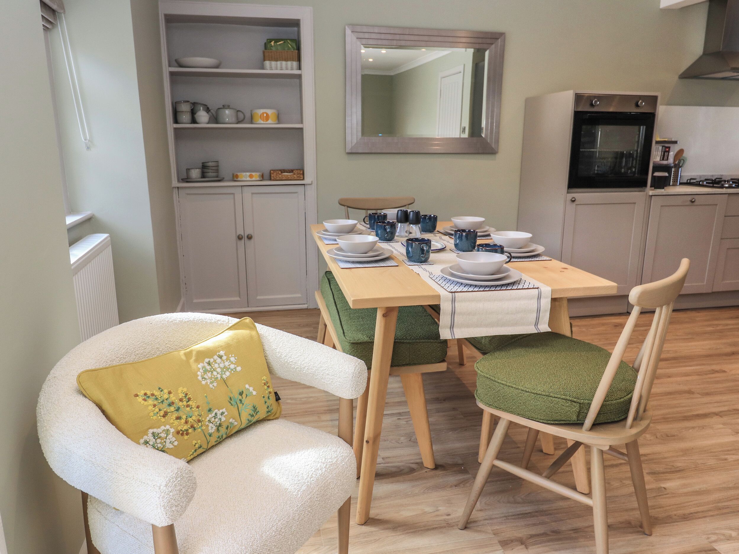 A dining area with a wooden table set for four, surrounded by chairs with green cushions. A white armchair with a yellow floral pillow is positioned in front of the table. A kitchen with a stove and oven is visible in the background.