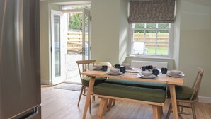 A dining area with a table set for four, chairs with green cushions, a window with a view of greenery, and a door leading outside. - 30 Northgate (Peebles)
