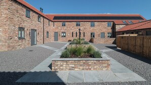 A brick building with a red-tiled roof, multiple windows, and a gravel courtyard with a stone planter. - The Stables (Thirsk)