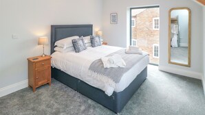 A bedroom with a bed, bedside tables, a mirror, and a view of a brick building through a window. - The Stables (Thirsk)