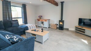 A living room with a sofa, a coffee table, a TV stand, a fireplace, and a window with curtains. - The Stables (Thirsk)