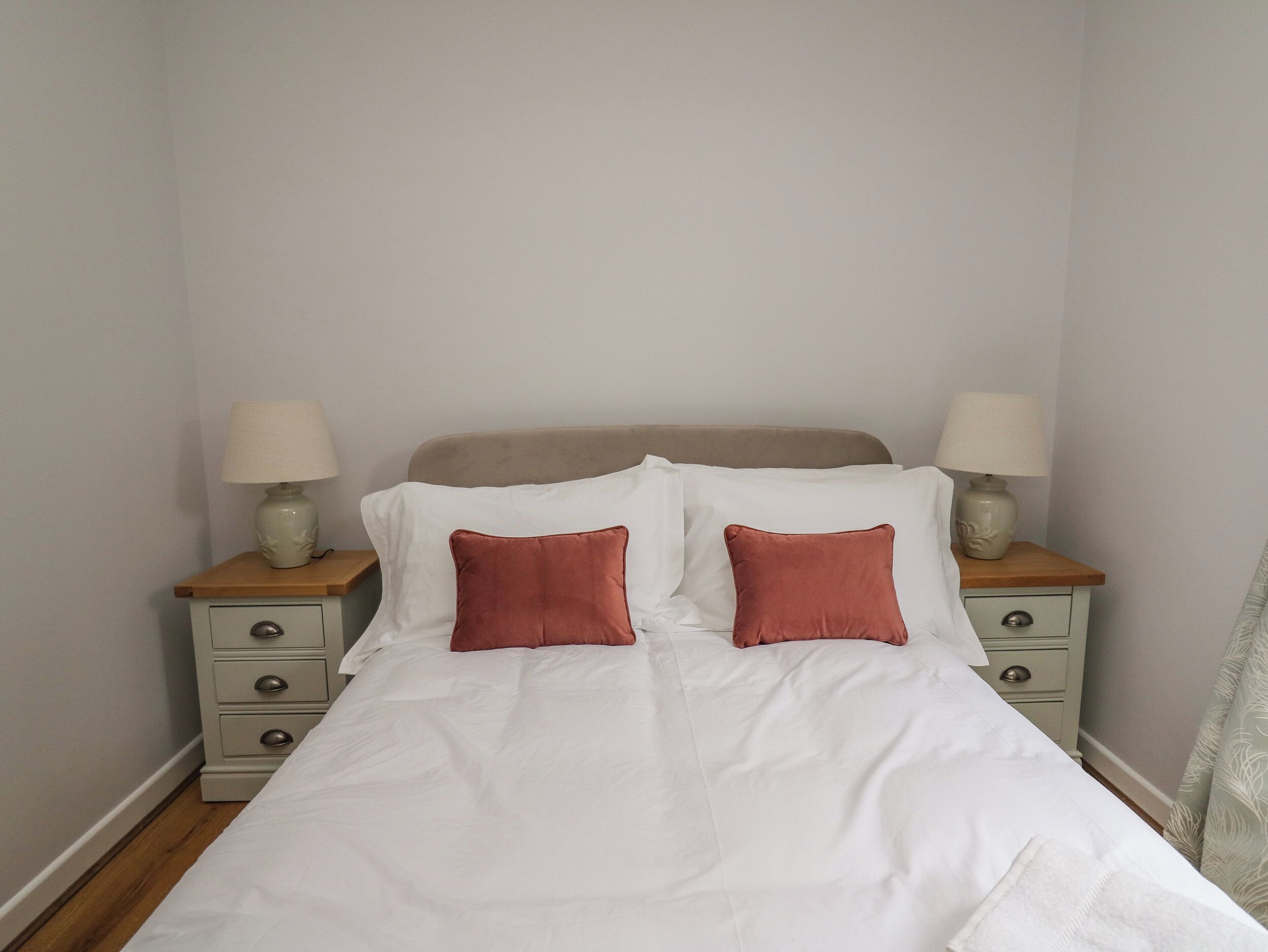 Una cama bien hecha con sábanas blancas y dos almohadas rojas, con dos mesitas de noche a cada lado, cada una con una lámpara y un cajón pequeño.