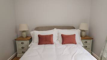 Una cama bien hecha con sábanas blancas y dos almohadas rojas, con dos mesitas de noche a cada lado, cada una con una lámpara y un cajón pequeño.