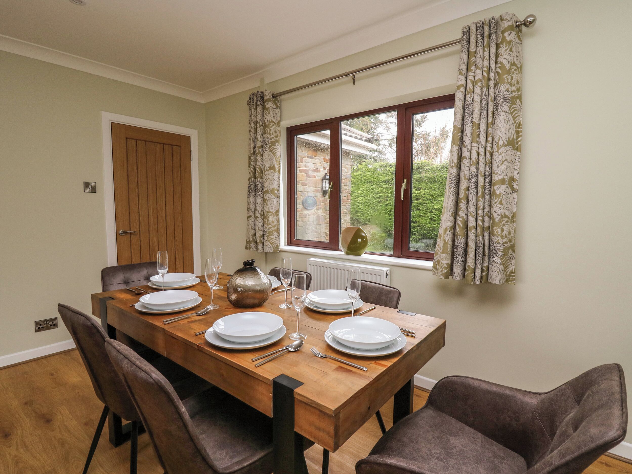 A dining room with a wooden table set for four, a window with curtains, and a door.