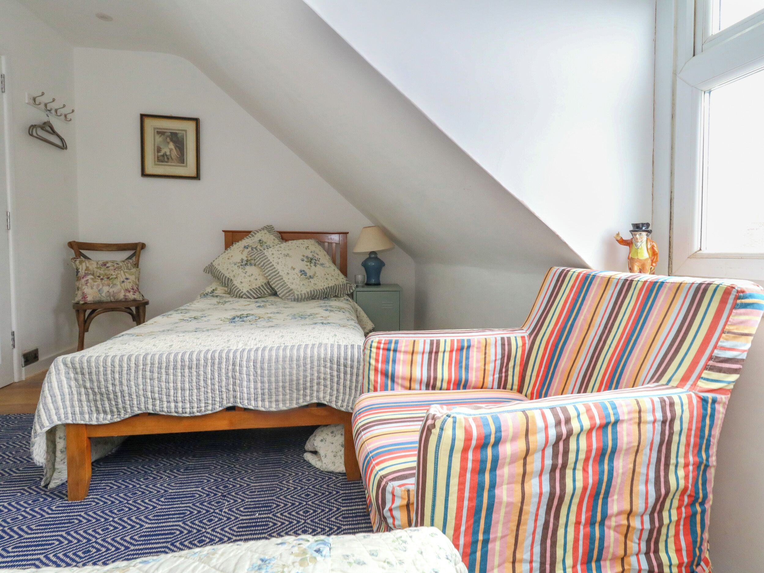 A bedroom with a sloped ceiling, a bed with floral bedding, a striped armchair, and a small table with a lamp.