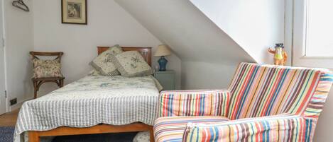 A bedroom with a sloped ceiling, a bed with floral bedding, a striped armchair, and a small table with a lamp.